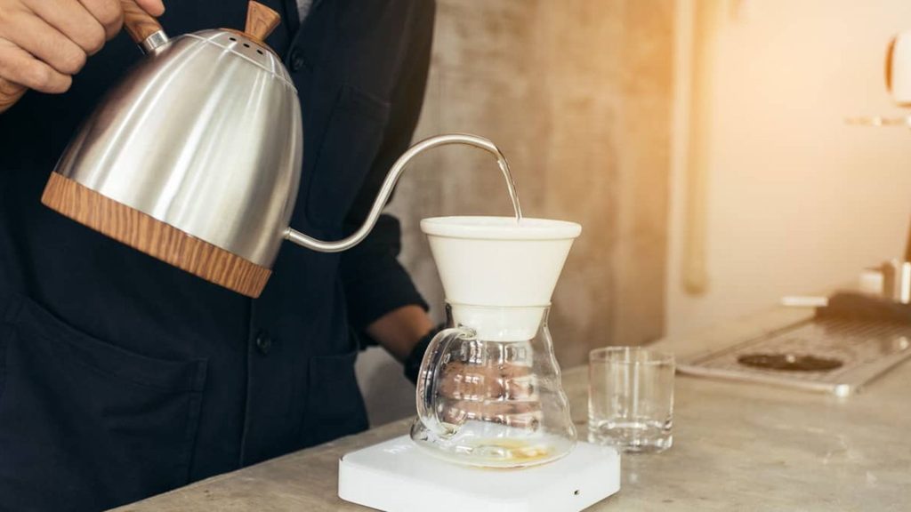 A person pours hot water from a gooseneck kettle into a pour-over coffee maker with a glass carafe on a scale, carefully preparing their coffee order on the countertop.