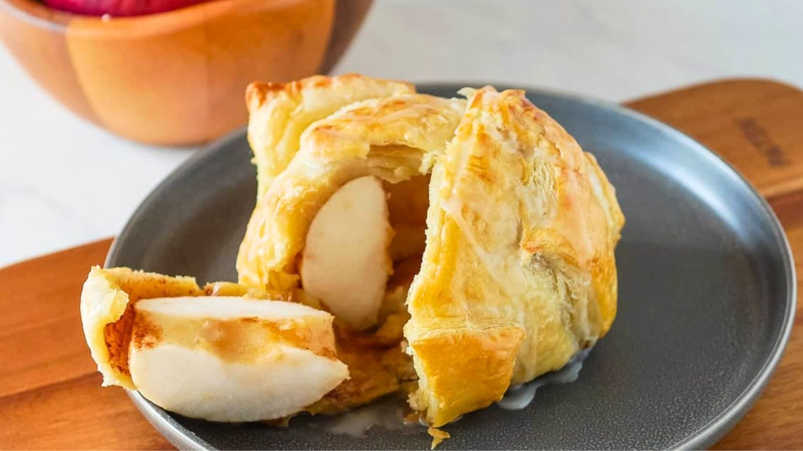 A baked apple wrapped in golden puff pastry is served on a gray plate, with a slice cut to show the apple filling inside.
