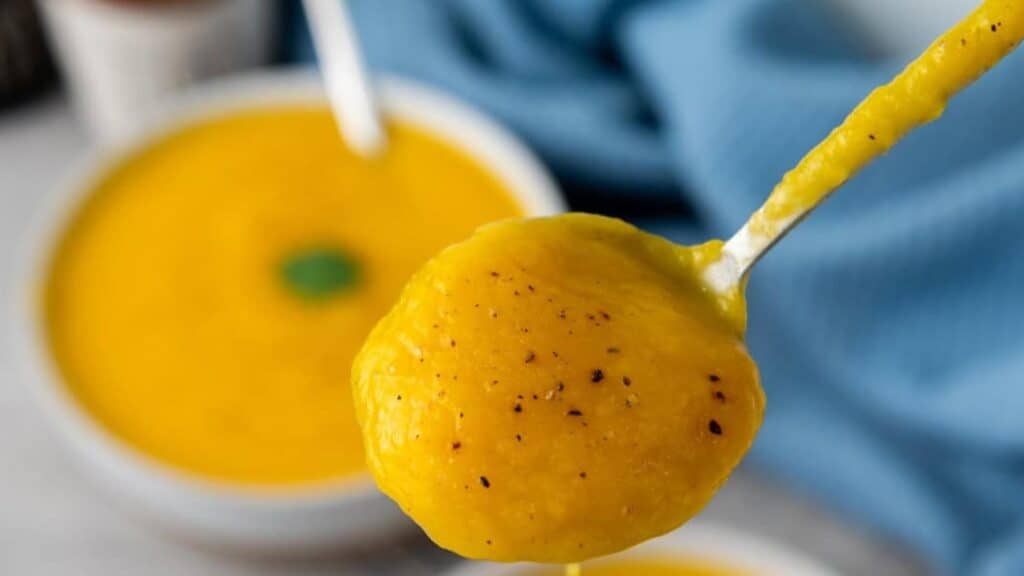 A spoonful of thick yellow soup with black pepper, held above a bowl of the same soup, with a blue cloth in the background.