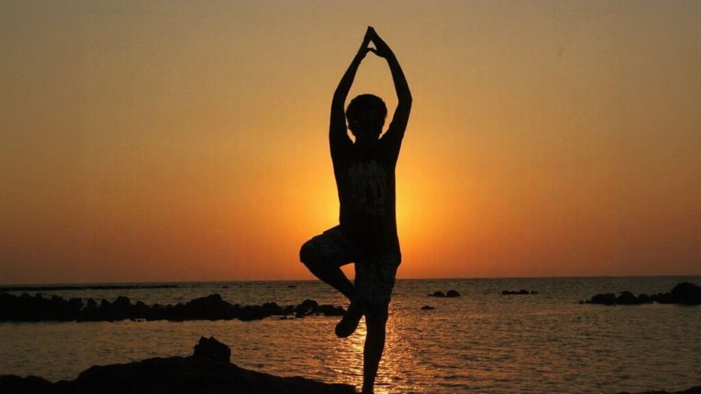 Silhouette of a person doing a yoga pose on one leg by the sea at sunset, with the sun low on the horizon and calm water in the background.