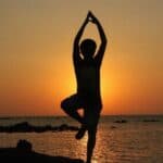 Silhouette of a person doing a yoga pose on one leg by the sea at sunset, with the sun low on the horizon and calm water in the background.