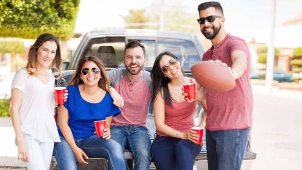 Five adults sit on the back of a pickup truck, smiling. Four hold red cups, and one holds a football toward the camera. It is daytime and they are outdoors.
