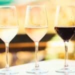 Three wine glasses filled with white, rosé, and red wine are arranged side by side on a table, ready for a Somm Smackdown, with a wine bottle partially visible on the right.