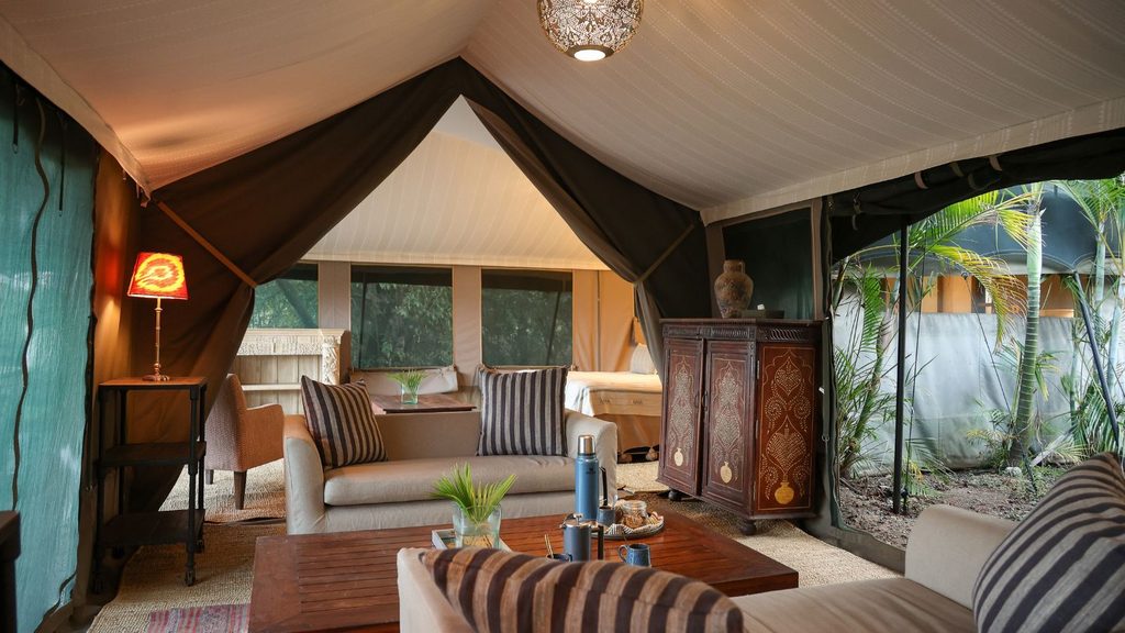 Luxury tent interior with sofas, a wooden table, a patterned cabinet, beds in the background, and greenery visible through mesh windows.