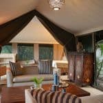 Luxury tent interior with sofas, a wooden table, a patterned cabinet, beds in the background, and greenery visible through mesh windows.