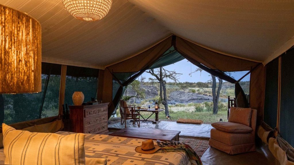 Interior of a luxury safari tent with a bed, dresser, lamp, and outdoor seating, overlooking a natural landscape with trees and rocks.