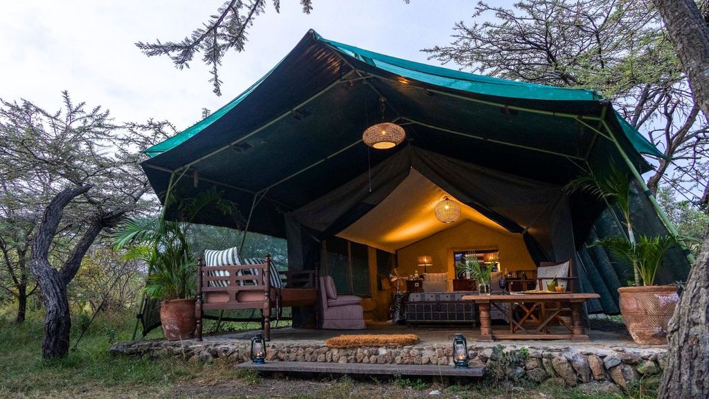 Large canvas safari tent with a shaded porch, outdoor furniture, lanterns, and potted plants, set in a wooded area with trees and greenery around.