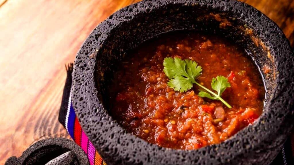 A stone molcajete filled with chunky red salsa, garnished with a sprig of cilantro, sits on a colorful striped cloth on a wooden surface.