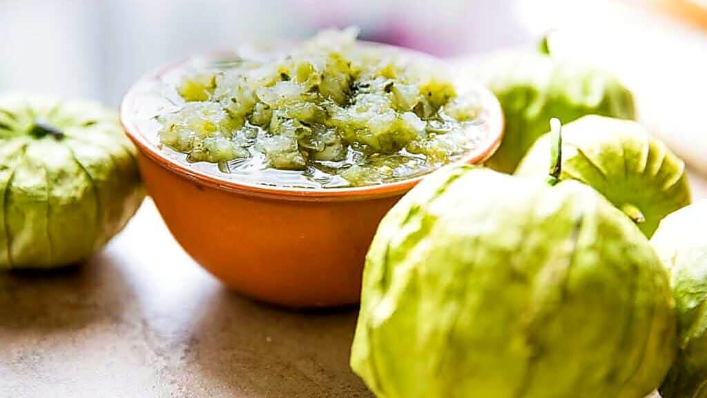 A bowl of green tomatillo salsa sits on a surface, surrounded by whole fresh tomatillos.