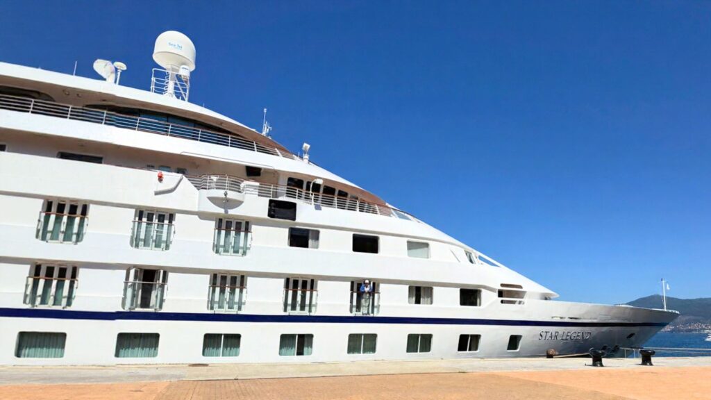 A white cruise ship docked at a port under a clear blue sky, showing multiple windows and antennas on the upper deck.