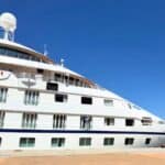 A white cruise ship docked at a port under a clear blue sky, showing multiple windows and antennas on the upper deck.