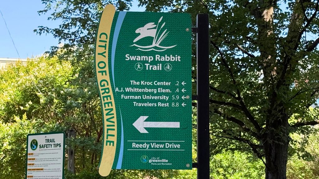 Green sign for the Swamp Rabbit Trail in Greenville, SC, lists distances to local destinations and points left to Reedy View Drive. Trees and a trail safety tips sign are in the background, providing helpful guidance for everyone enjoying the Swamp Rabbit Trail.