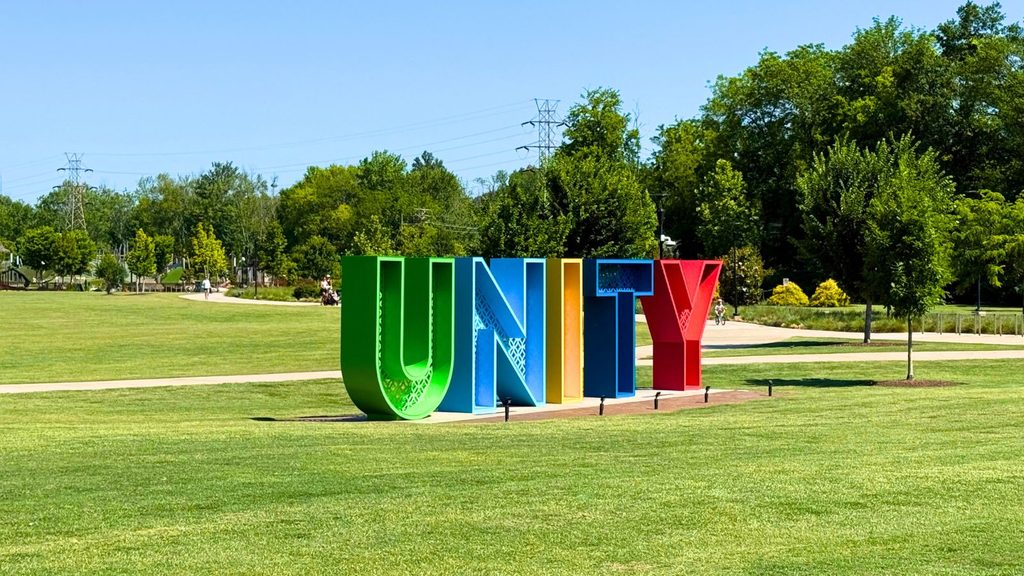 Large colorful letters spelling &ldquo;UNITY&rdquo; stand on a grassy field in a park along the scenic Swamp Rabbit Trail, with trees and a clear blue sky in the background.