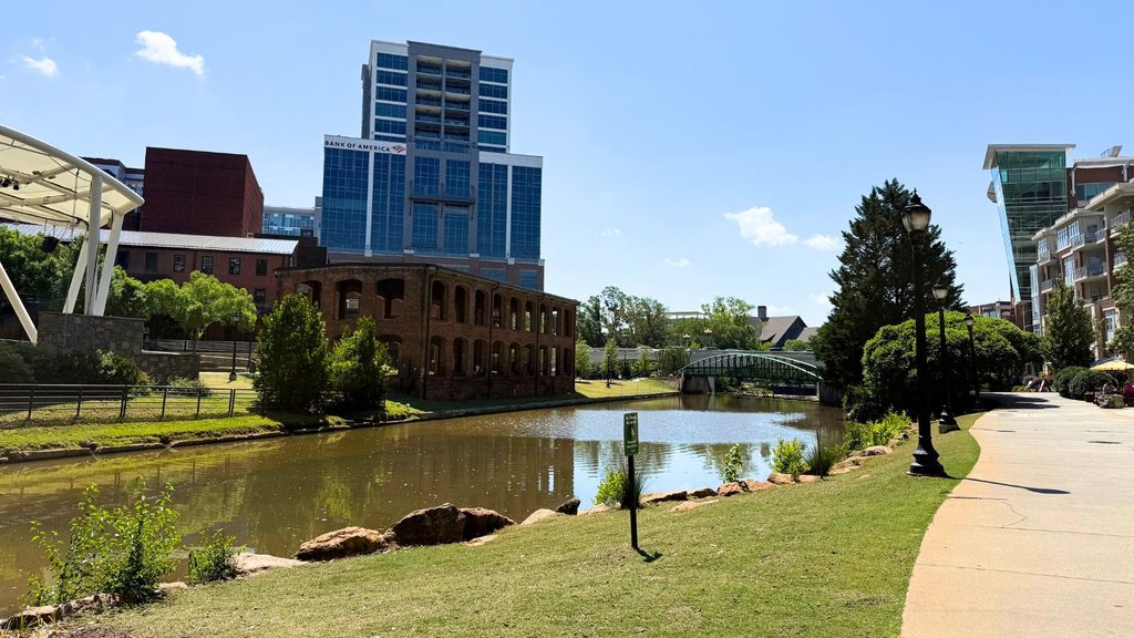 A paved walkway runs beside a calm river along the Swamp Rabbit Trail, with a brick building, modern high-rise, and greenery visible under a clear blue sky.