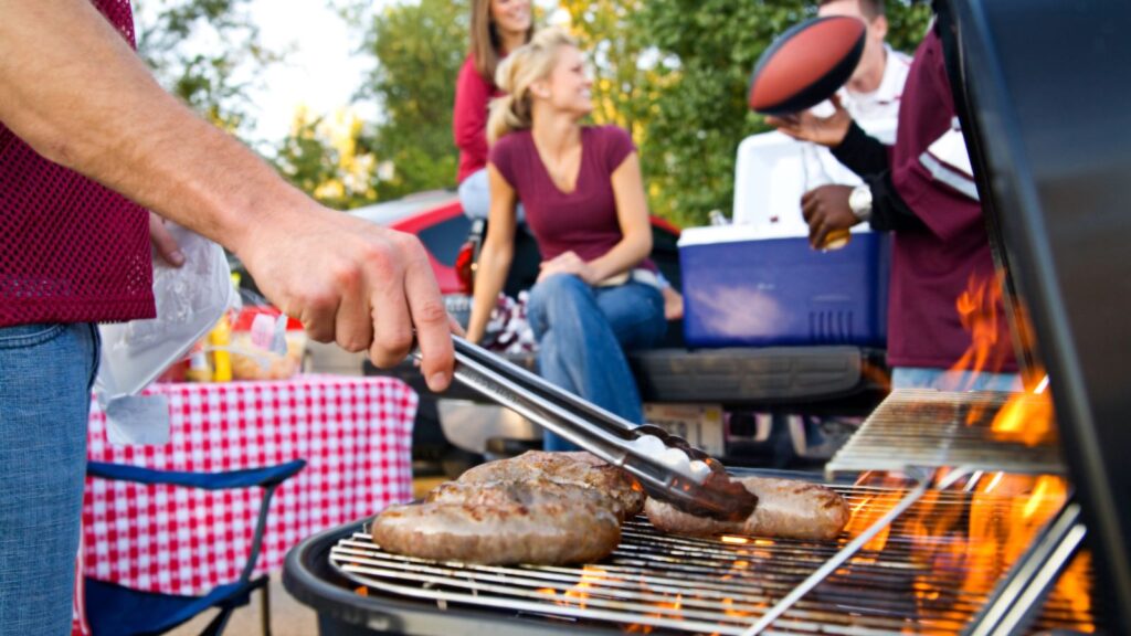 Person grilling meat on a barbecue at an outdoor gathering, with people in the background near a table and a cooler.
