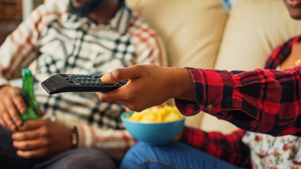 Two people sit on a couch. One person holds a TV remote, while the other holds a green bottle. A blue bowl of snacks is on their lap. Both wear plaid shirts.