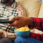 Two people sit on a couch. One person holds a TV remote, while the other holds a green bottle. A blue bowl of snacks is on their lap. Both wear plaid shirts.