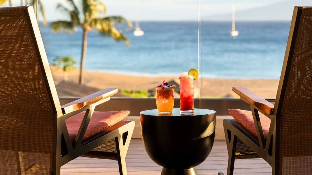 Two cocktails sit on a small table between two chairs on a balcony overlooking a sandy beach and ocean, with palm trees and anchored boats in the background.