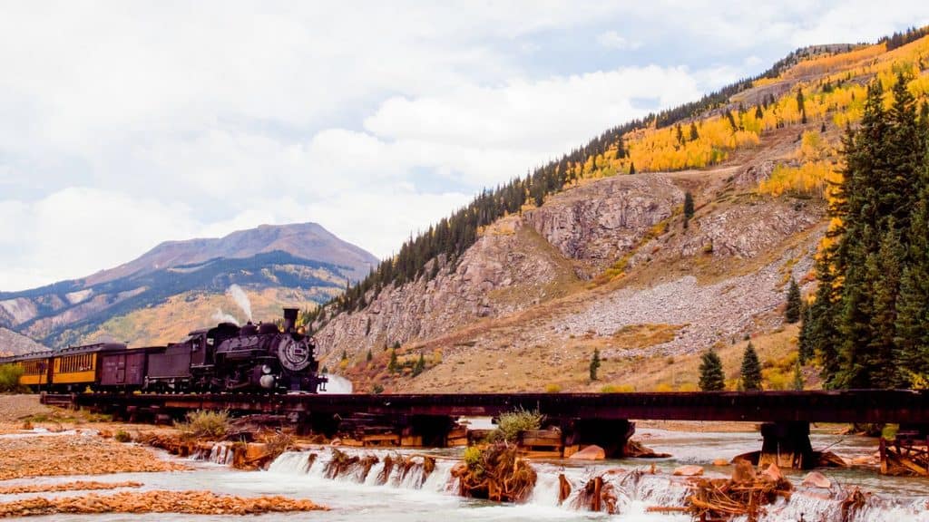 A black steam train crosses a rust-colored bridge over a small river, with mountains and autumn trees in the background&mdash;an idyllic scene perfect for fall train rides.