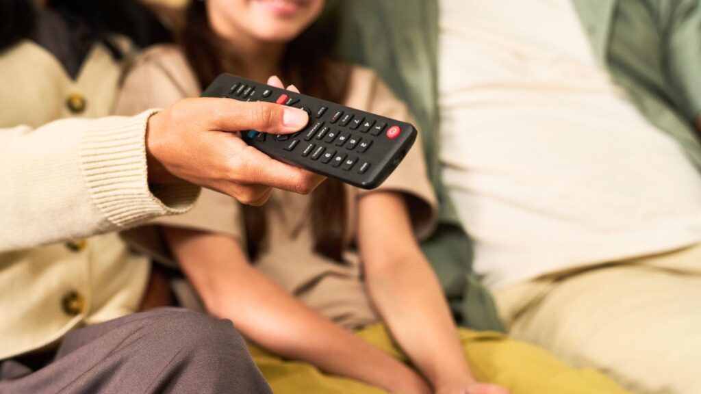 A person holds a TV remote control, with two others seated beside them, partially visible in the background.