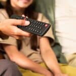 A person holds a TV remote control, with two others seated beside them, partially visible in the background.