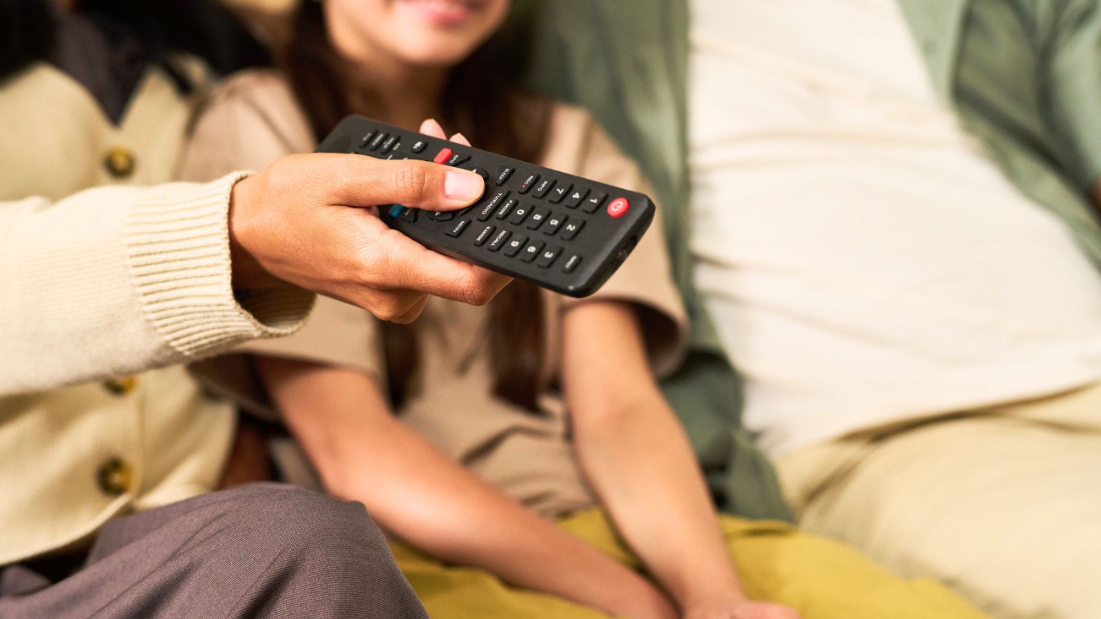 A person holds a TV remote control, with two others seated beside them, partially visible in the background.
