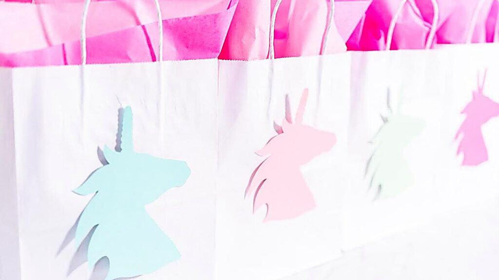 White gift bags with pink tissue paper, each decorated with a pastel-colored unicorn silhouette cutout.