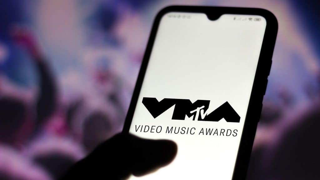 A hand holds a smartphone displaying the MTV Video Music Awards (MTV VMAs) logo on a white screen, with a blurred, colorful background.