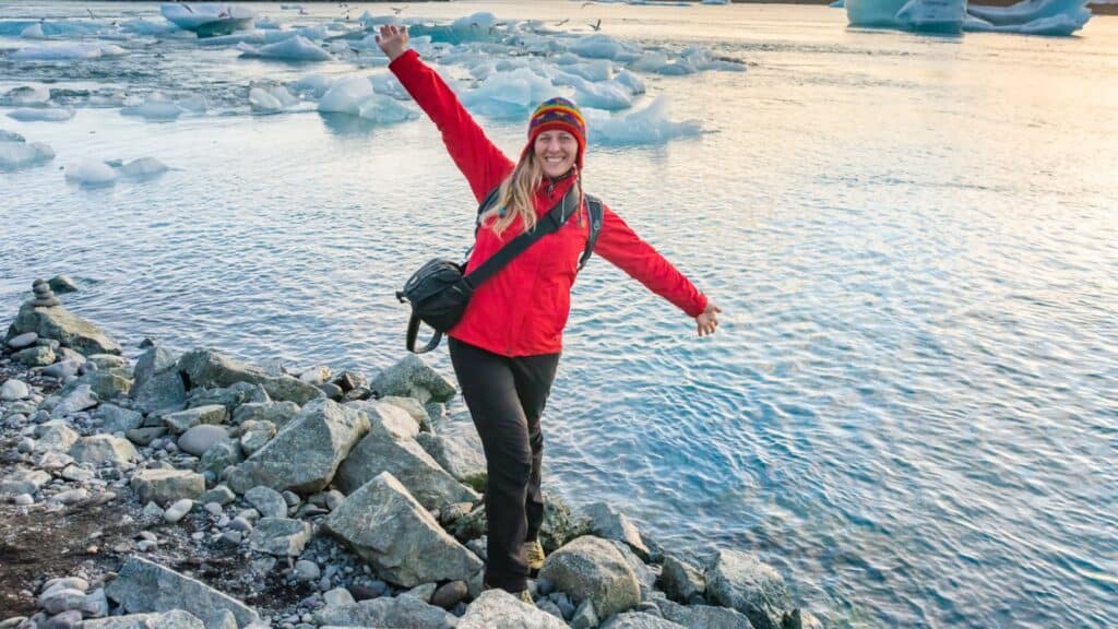Person in a red jacket stands on rocky shore with one arm raised, smiling at the camera; icy water and floating ice in the background.