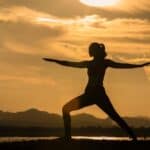 A person practices yoga in a warrior pose outdoors at sunset, with mountains and clouds visible in the background.