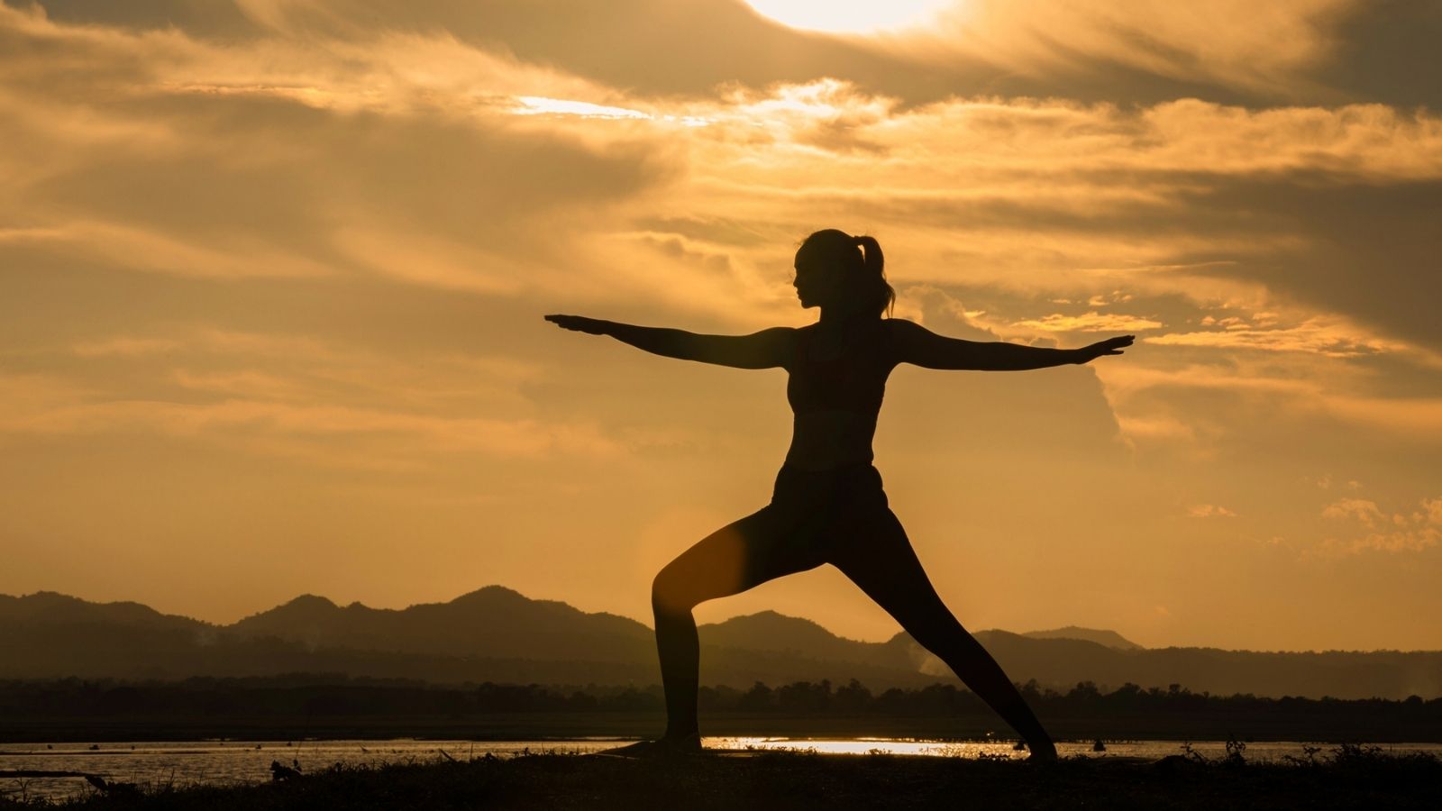 A person practices yoga in a warrior pose outdoors at sunset, with mountains and clouds visible in the background.