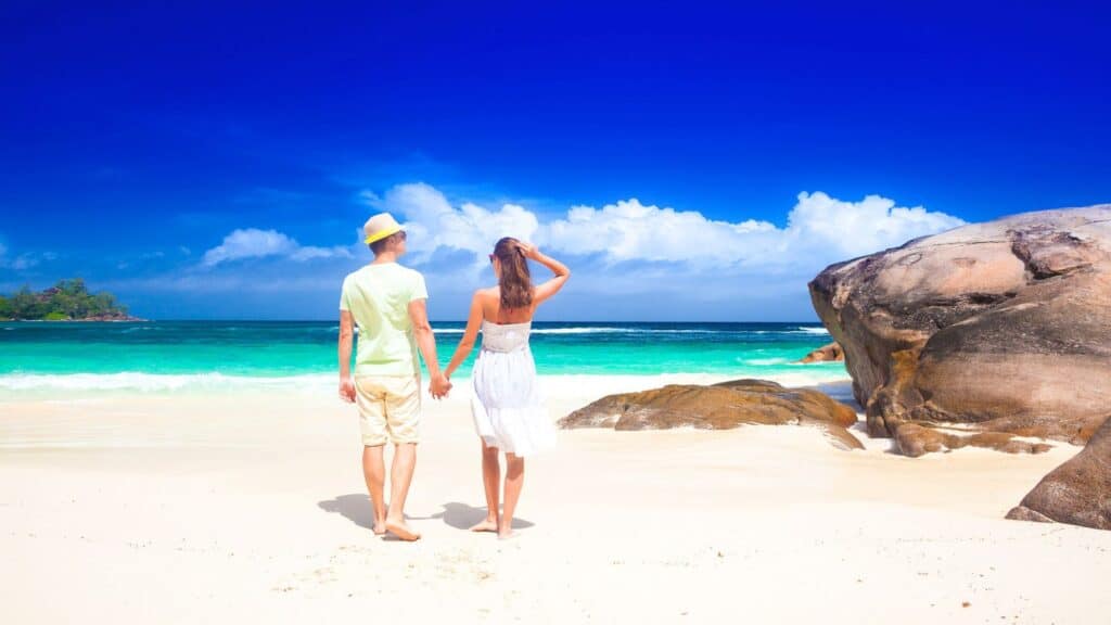 A man and woman holding hands stand on a sandy beach, facing the turquoise sea and blue sky, with rocks on the right side—an idyllic scene perfect for those seeking post-Labor Day resort deals.