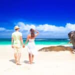 A man and woman holding hands stand on a sandy beach, facing the turquoise sea and blue sky, with rocks on the right side—an idyllic scene perfect for those seeking post-Labor Day resort deals.