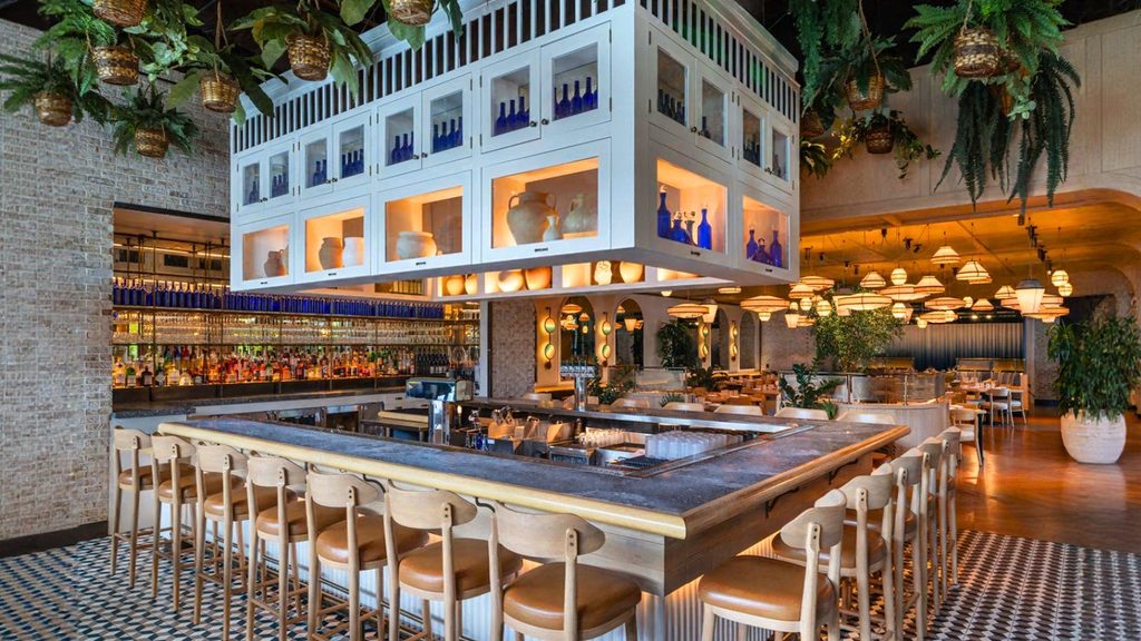 Modern restaurant interior with a central bar surrounded by wooden chairs, hanging plants above, and decorative lighting throughout the space.