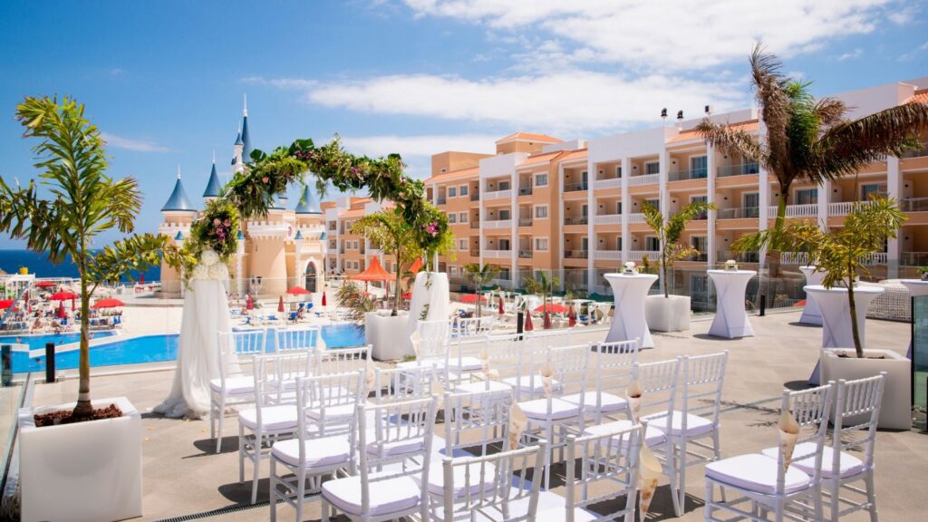 Rows of white chairs set up for a Caribbean wedding packages ceremony on a terrace overlooking a pool and resort.