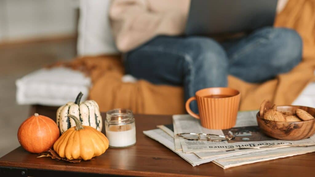 A coffee table with mini pumpkins, a lit candle, a mug, cookies, and a newspaper, with a person on a sofa; part of our fall-themed hotel packages.