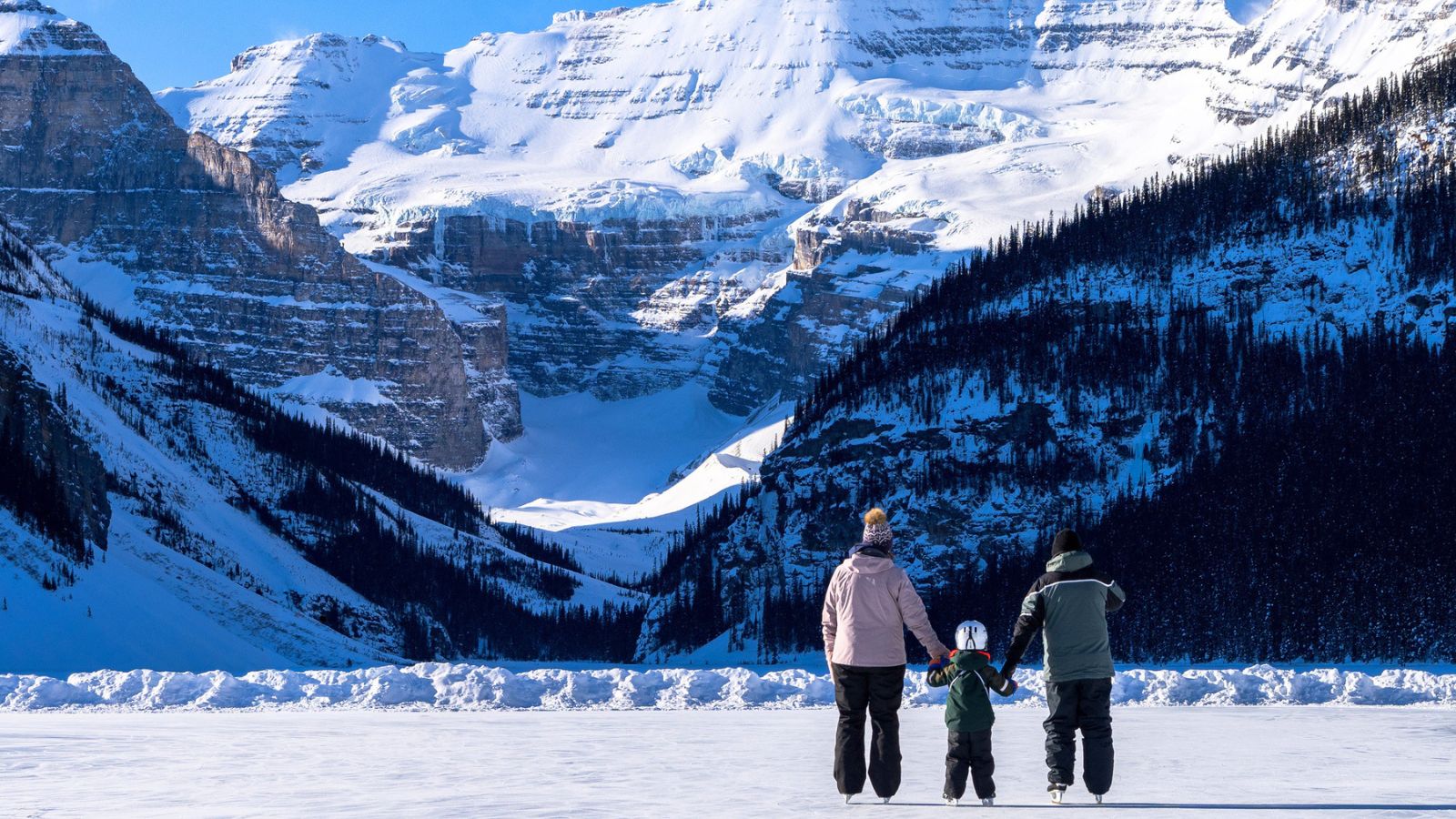 A family of three enjoys their family vacation ice skating on a frozen lake in front of a snowy mountain range.