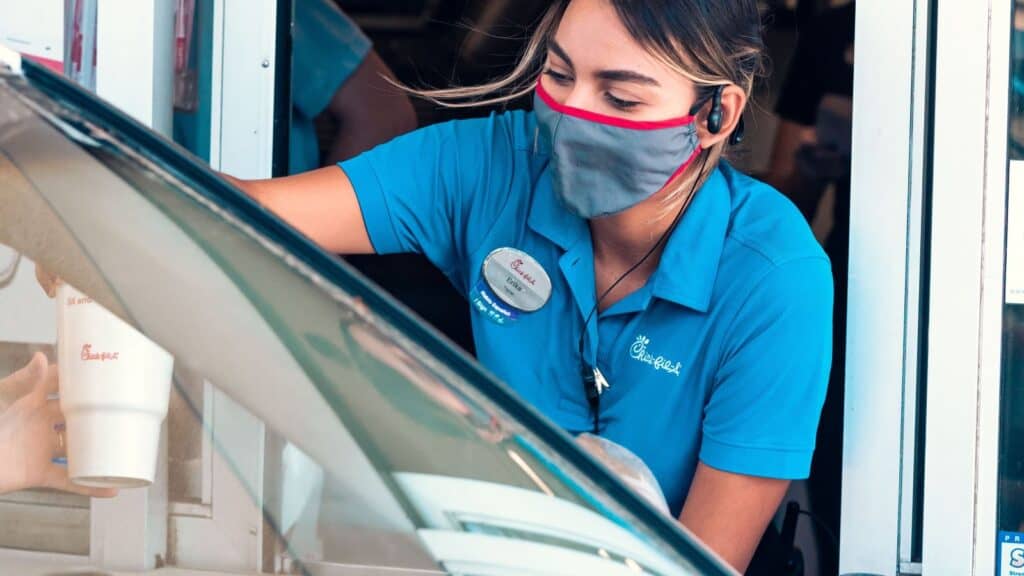 A Chick-fil-A employee in a blue uniform and face mask hands a drink through a drive-thru window, a modern evolution of the invention of the drive-thru.