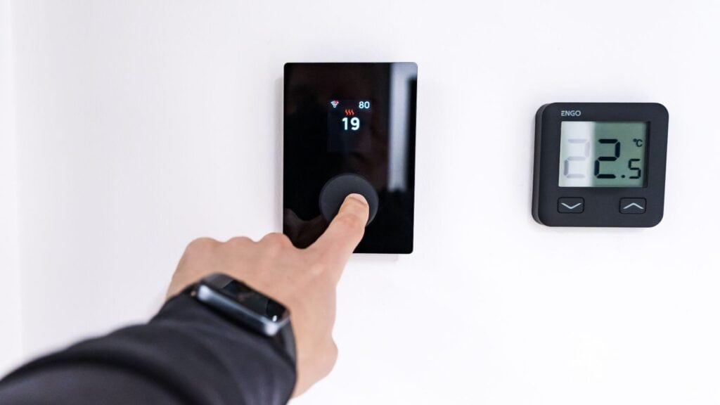 A finger presses the dial on a black smart thermostat used to weatherproof a home before a cold snap, mounted next to an Engo digital display showing 22.5 degrees Celsius.