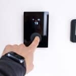A finger presses the dial on a black smart thermostat used to weatherproof a home before a cold snap, mounted next to an Engo digital display showing 22.5 degrees Celsius.