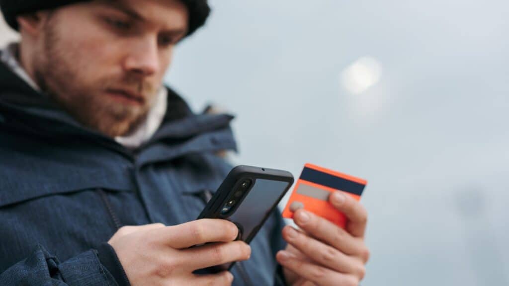 A man in a winter jacket holds a smartphone and a credit card while potentially falling for a vacation rental scam.