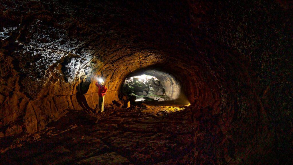 A person with a headlamp stands in a dark volcanic lava tube that feels like a set for scary movies, looking toward an opening that reveals greenery and light.