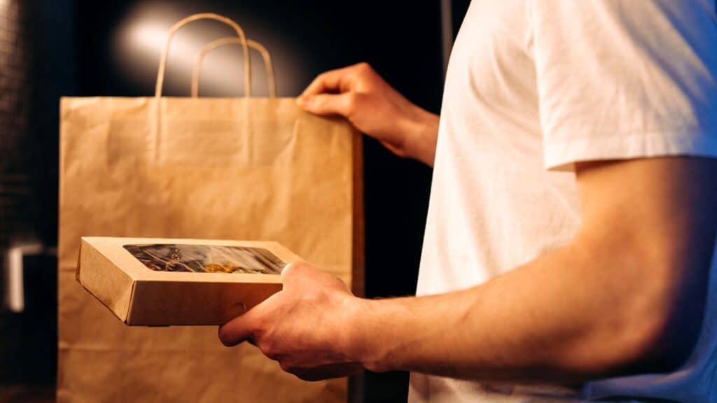 A person in a white t-shirt holding a food takeout box in front of a paper bag as home cooks legally sell food.