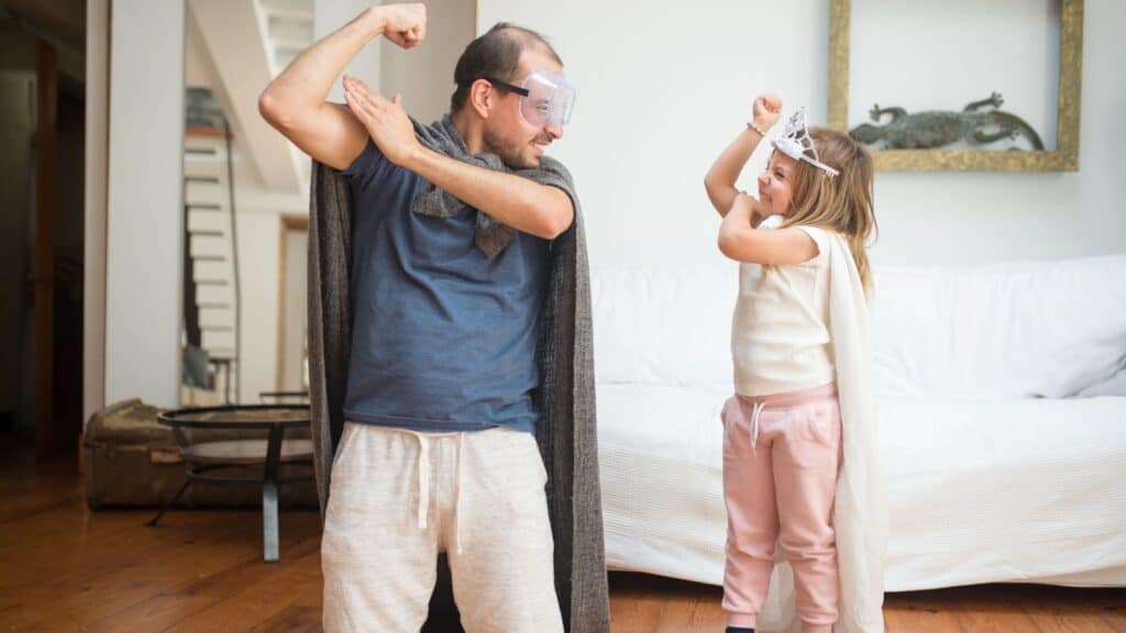 A man wearing a cape and goggles and a young girl wearing a cape and tiara pose like superheroes in family Halloween costumes at home.