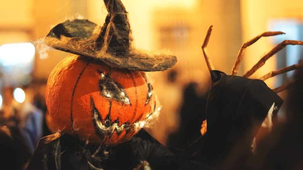 A person with long, claw-like fingers wearing a witch&rsquo;s hat and a jack-o&rsquo;-lantern mask, typical of theme parks with Halloween events.