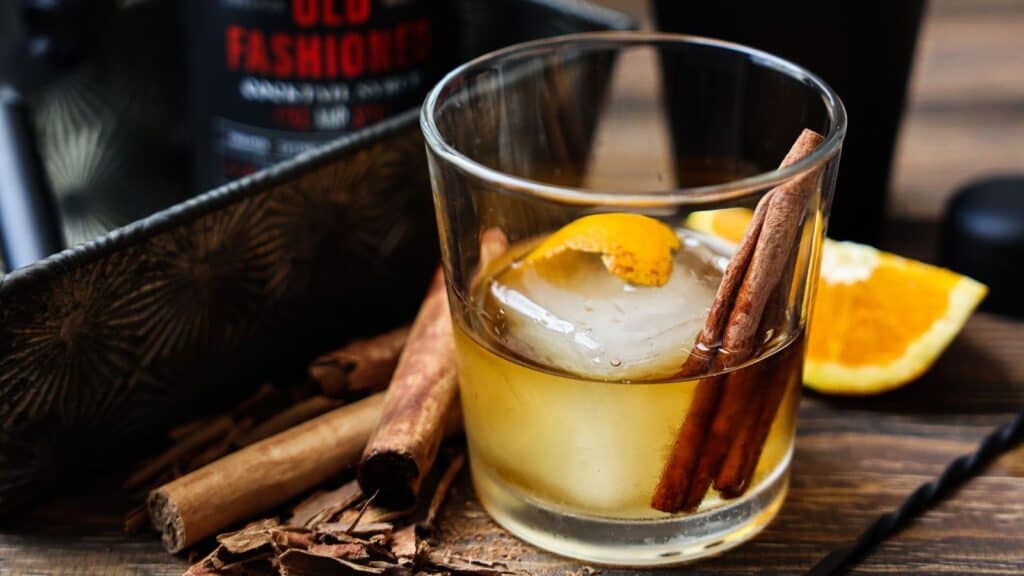 An Old Fashioned Halloween cocktail with a large ice cube, orange peel, and cinnamon stick garnish, set on a wooden table with loose cinnamon sticks and orange slices.