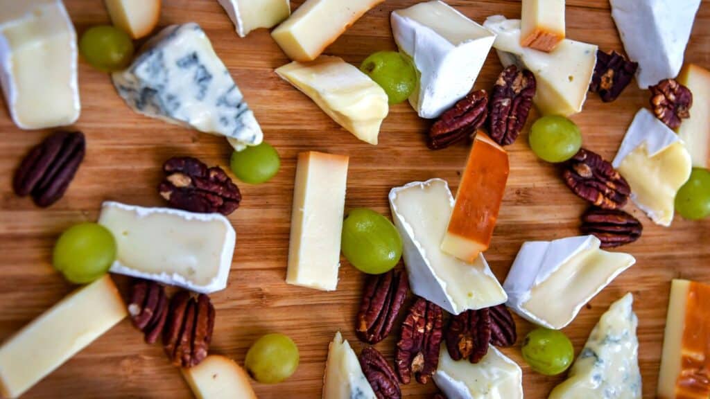 A variety of cheese wedges, green grapes, and pecan halves arranged on a wooden board for seasonal fall cheese pairings.