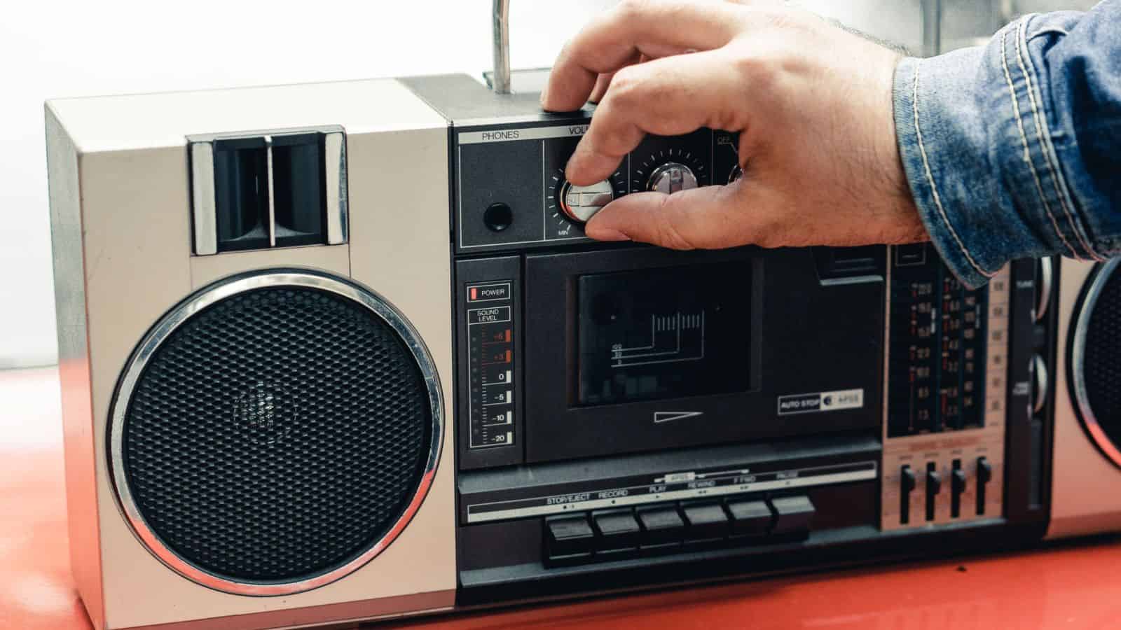 A hand adjusting the volume knob on a vintage portable cassette player, evoking boom box memories.