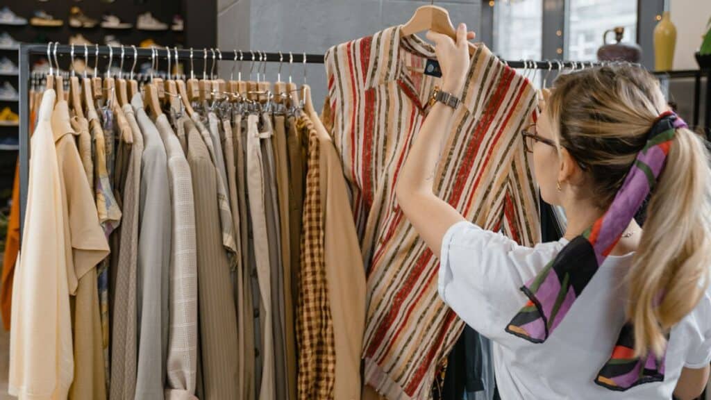 A person browses a rack of clothing for QVC and HSN impulse buys, holding up a red and tan striped button-down shirt.