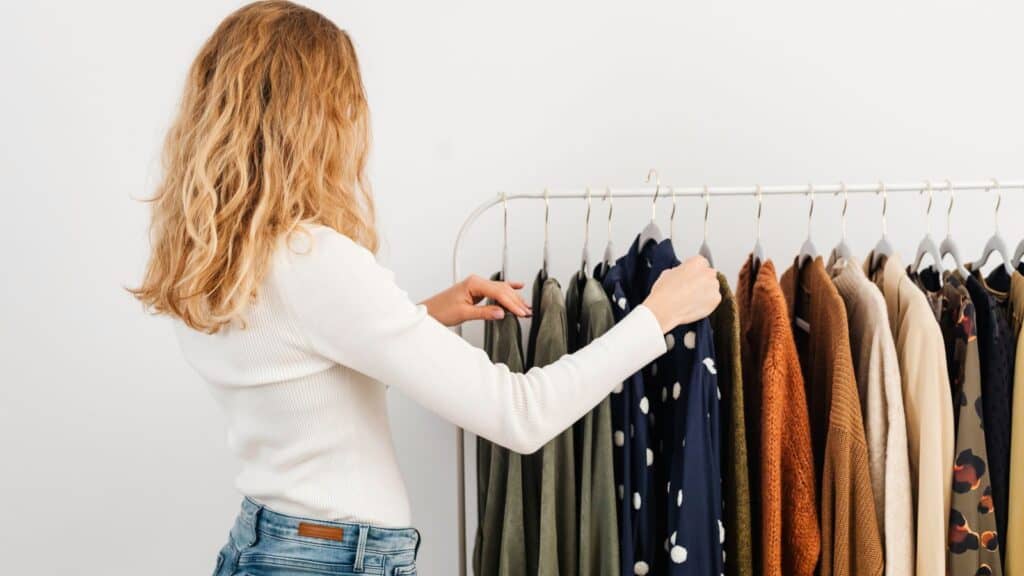 A woman in a white shirt browsing a clothing rack of shirts and sweaters while avoiding fall closet-swap mistakes.